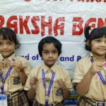 Three schoolgirls in beige uniforms display handmade crafts in front of a banner at a school event